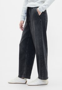 Črne široke kavbojke z žepi ob straneh, elastičnim pasom z vrvico za zategovanje in ohlapnim krojem. Oblečene z belimi športnimi copati.