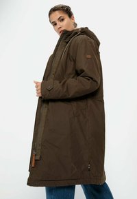 Braune, knielange Parka mit Kapuze, ausgestattet mit Reißverschluss und Druckknopfverschlüssen, seitlichen Taschen und einer strukturierten Oberfläche. Akzentuiert mit Lederdetails.