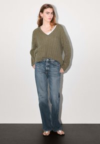 Olive green cable-knit sweater with V-neck, paired with faded blue flared jeans. Model stands on a dark floor against a light background.