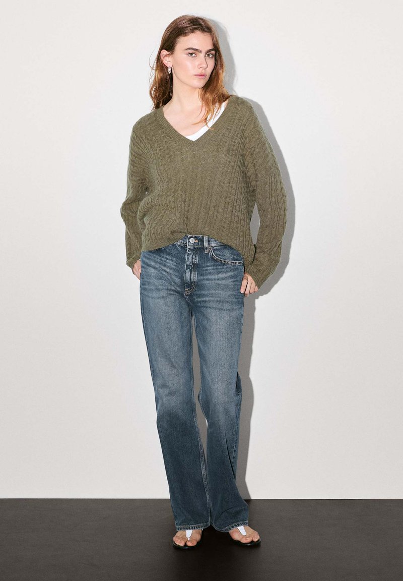 Olive green cable-knit sweater with V-neck, paired with faded blue flared jeans. Model stands on a dark floor against a light background.