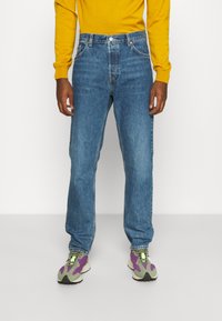 Vaqueros azul denim de corte recto, con cinco bolsillos, bragueta con botones y un sutil desgaste. Combinados con un suéter amarillo y zapatillas deportivas coloridas.