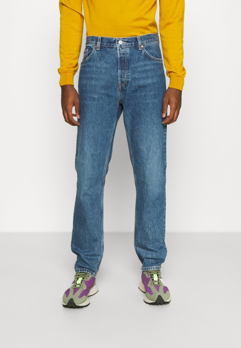 Vaqueros azul denim de corte recto, con cinco bolsillos, bragueta con botones y un sutil desgaste. Combinados con un suéter amarillo y zapatillas deportivas coloridas.