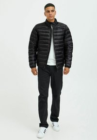 Svart polstret jakke med høy krage, med glidelås foran og horisontal quilted design. Båret med en hvit skjorte, svarte bukser og joggesko.