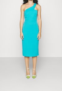 Une robe ajustée, à une épaule, en tissu turquoise vif, arrivant au genou, assortie à des chaussures à talons hauts vertes ornées de strass.