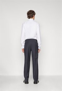 Witte lange mouwen shirt met knoopmanchetten, gecombineerd met marineblauwe broek, zichtbare achterzakken en zwarte schoenen, gezien van achteren.