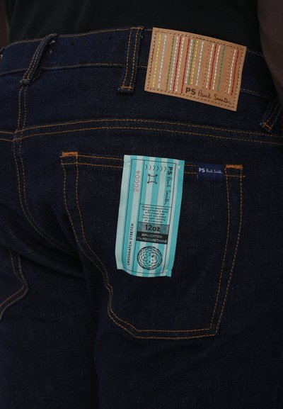 Jeans de mezclilla oscura con costuras naranjas; presenta un parche de cuero marrón con rayas coloridas y una etiqueta de cuidado turquesa en el bolsillo trasero.