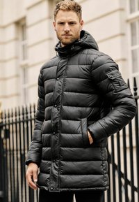 Sort pufferjakke med hætte, der har et quiltet design, trykknapper, sidelommer og et patch på venstre ærme. Glat tekstur.