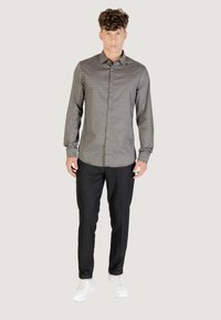Giovane che indossa una camicia grigia a maniche lunghe con motivo, pantaloni neri aderenti e sneakers bianche, in piedi contro uno sfondo neutro.