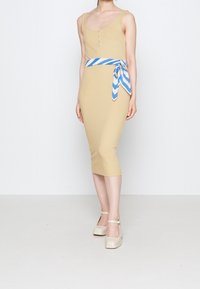 Přiléhavé ribové midi šaty v béžové barvě s kulatým výstřihem, knoflíky a páskem s modro-bílými pruhy. V kombinaci s béžovými podpatky se zapínáním kolem kotníku.