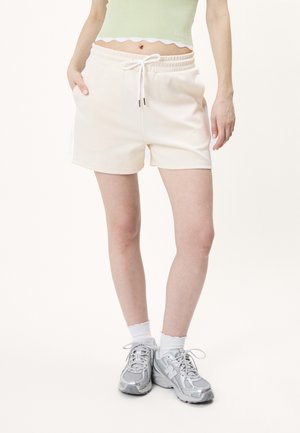 Frau trägt hellbeige Shorts mit Kordelzug, weiße Socken und silbergraue Sportschuhe und steht mit einer Hand in der Tasche.