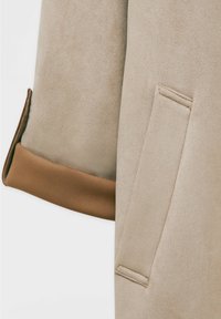 Manche de veste en daim beige avec un revers brun plié et un détail de poche verticale cousue sur un fond clair.