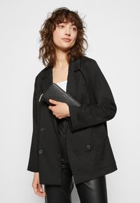 Blazer noir à double boutonnage avec de grands boutons, porté sur un haut blanc. Le modèle tient un portefeuille en cuir noir, avec une fermeture à zip.