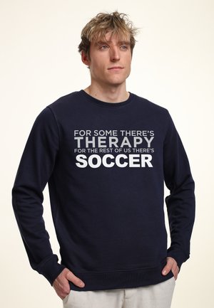 Námorně modrá mikina s kulatým výstřihem a dlouhými rukávy. Bílý a šedý text zní: "PRO NĚKTERÉ EXISTUJE TERAPIE, PRO OSTATNÍ ZAS FOOTBALL."