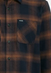 Plaid shirt in dark blue and brown, featuring a button-up front, two chest pockets, and a Volcom logo label on the left pocket.