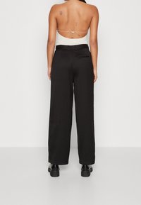 Žena v černých širokých kalhotách, krémovém topu bez zad s tenkým ramínkem, černých botách, stojící zády k fotoaparátu na bílém pozadí.