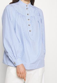 Blusa a righe blu e bianche con colletto alto, pattina con bottoni e dettagli arricciati sul giromanica. Maniche a palloncino con bottoni sui polsini.