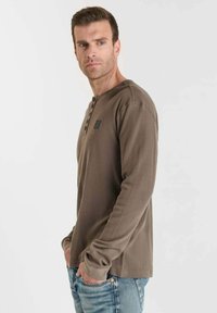 Camisa de manga larga de color marrón oscuro con una tela texturizada, cuello redondeado y tres botones. Detalle del logo sutil en el lado izquierdo del pecho.