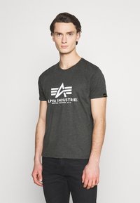 Tmavě šedé tričko s krátkými rukávy má bílé logo se nápisem "ALPHA INDUSTRIES" a malou etiketou na rukávu. Hladká textura.