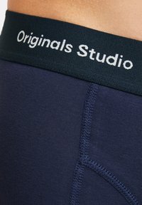 Dunkelblaue Fahrradhosen mit einem breiten schwarzen Bund, der mit weißer Schrift "Originals Studio" gekennzeichnet ist. Die Hose besteht aus glattem elastischem Material und hat flache Nähte.