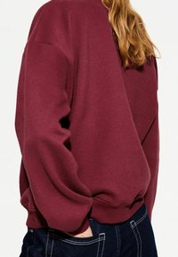 Vinröd sweatshirt med avslappnad passform, ribbad fåll och muddar, med en mjuk textur. Kombinerad med mörka denimjeans.