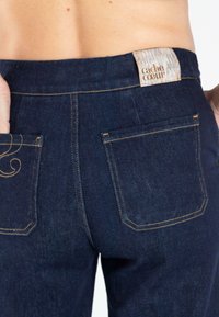Donkerblauwe denim jeans met achterzakken en een subtiele gouden stiksel detail. Heeft een merklabel op de tailleband "cache coeu" in licht hout.