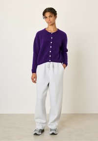 Ljubičasti puhasti kardigan s okruglim izrezom i prednjim gumbima u kombinaciji s svijetlosivim trenirka hlačama i sivim tenisicama. Jednostavan, ležeran kroj.