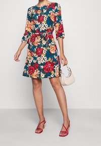 Blumenkleid mit 3/4-Ärmeln, tailliertem Bund, türkisfarbenem Hintergrund, roten und orangefarbenen Blüten. Model hält eine helle Handtasche und trägt rote High Heels.