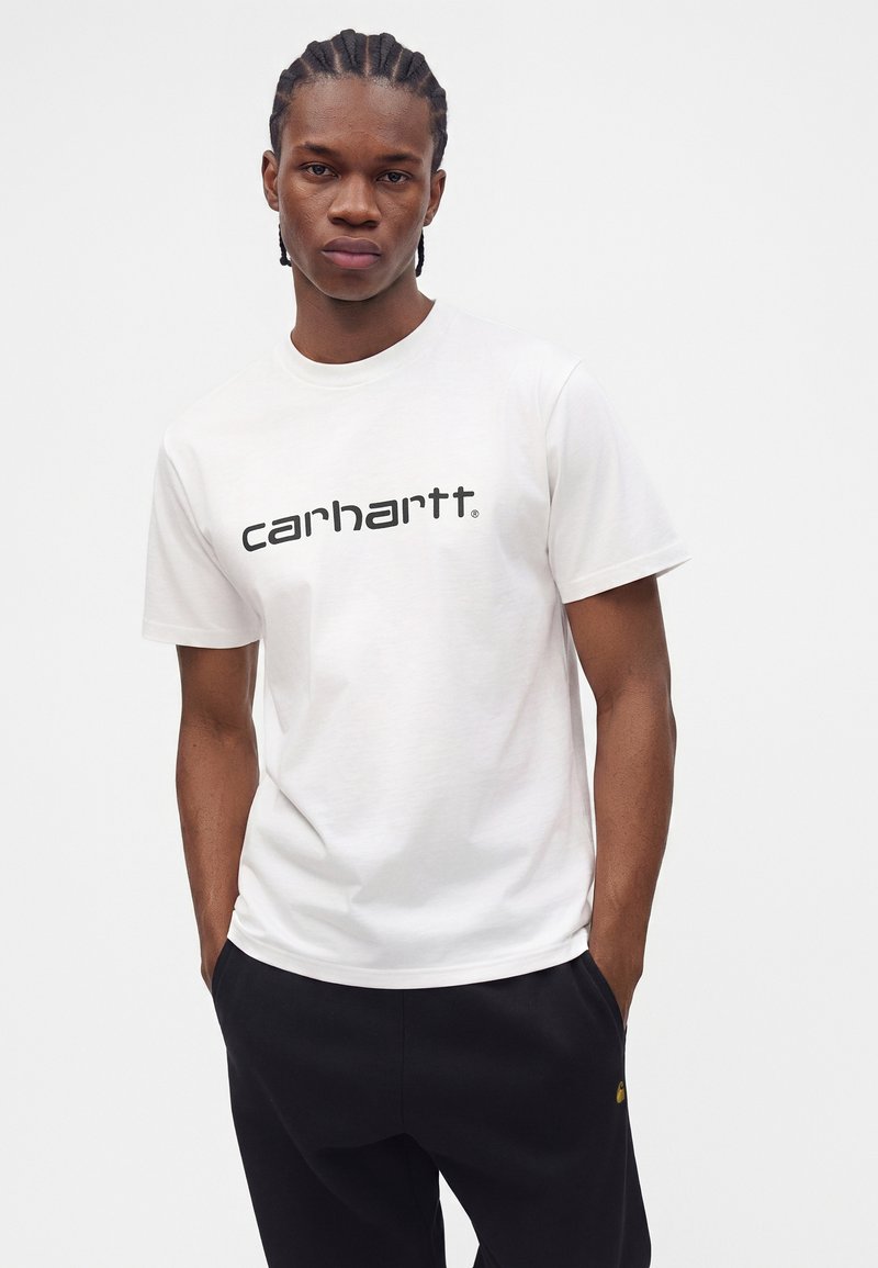 Young man with braided hair wearing a white Carhartt t-shirt and black pants with hands in pockets against a plain background.