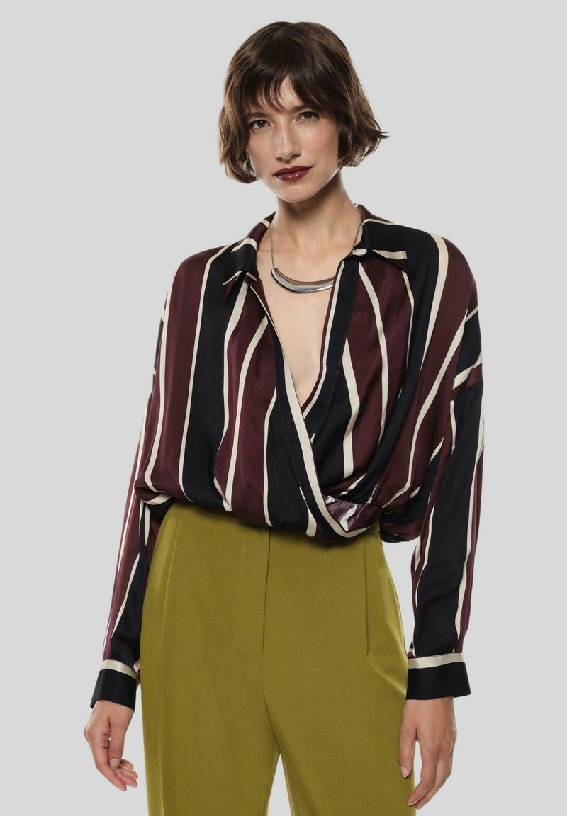 Blusa a maniche lunghe color bordeaux e nera con righe verticali, che presenta un profondo scollo a V e un tessuto morbido e liscio; abbinata a pantaloni verde oliva.