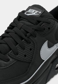 Sneaker nera Nike caratterizzata da una parte superiore in sintetico testurizzato, lacci neri piatti, accento swoosh grigio e una linguetta imbottita con logo.