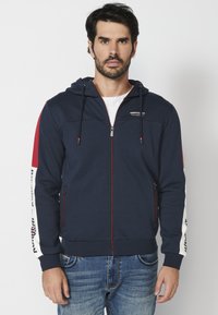 Tamsiai mėlyna zip-up gobtuvinė striukė su raudonomis ir baltomis šoninėmis juostelėmis, elastingomis rankovėmis ir raišteliu gobtyje. Savybės: logotipo detalė krūtinėje ir užtrauktukai ant kišenių.