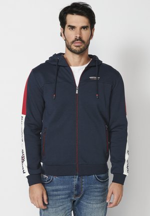 Marine blauwe zip-up hoodie met rode en witte zijstrepen, elastische manchetten en een trekkoord capuchon. Bevat een logo op de borst en ritsen op de zakken.