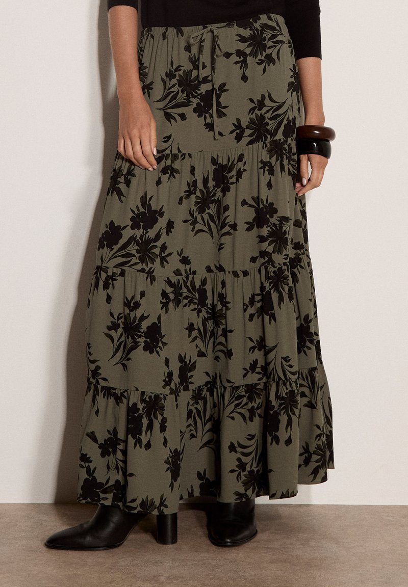 Longue jupe vert olive avec imprimé floral noir portée avec des bottes noires, un haut à manches longues noir et des bracelets larges foncés à un poignet.