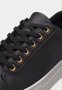 Κοντινή λήψη μαύρου παπουτσιού σνίκερ με μαύρα κορδόνια περασμένα από έξι χρυσά οπές, με απαλό δέρμα και λευκή λαστιχένια σόλα.