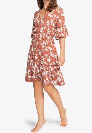 Femme portant une robe fleurie couleur rouille longueur genoux avec un ourlet et des manches volantés, debout pieds nus sur un fond blanc.