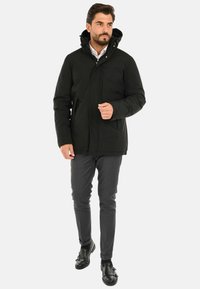 Giacca isolata nera con cappuccio, dotata di una cerniera frontale, tasche laterali e un design leggermente affusolato. Indossata sopra pantaloni grigi e scarpe nere.