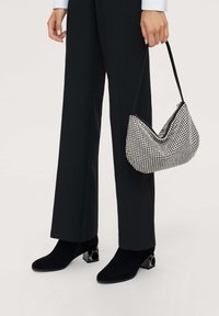 Bolso de mano de cuadros en blanco y negro con forma curva, hecho de un material texturizado, sostenido por una persona que viste pantalones anchos negros y botas de tacón.
