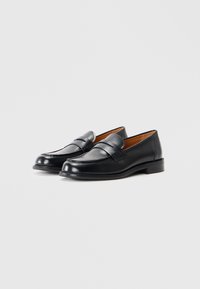 Mustad nahast loafersid sileda viimistlusega, dekoratiivne rihm ees ja madal ladustatud konts. Siseosa näitab pruuni voodrit.