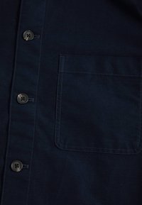 Navyblauwe shirt gemaakt van een gladde stof, met een knoopsluiting, vier knopen aan de voorkant en een gestikte zak aan de linkerkant.