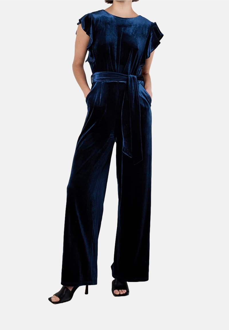 Femme portant une combinaison sans manches en velours bleu marine avec une ceinture nouée à la taille et un pantalon à jambes larges, associée à des talons ouverts noirs.