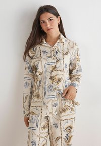Chemise couleur crème avec des motifs floraux et géométriques en bleu et marron. Avec un col, une fermeture à boutons et des manches longues.