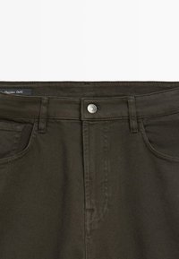 Donkergroen olijfgroene denim jeans taille met een sluiting aan de voorkant met knopen, riemlussen en voorzakken, met zichtbare stoftextuur en stikseldetails.