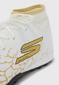 Witte sportieve schoen met een hoge schacht, met een gestructureerd oppervlak, een goudkleurig gebarsten patroon en een zwarte zool met noppen.