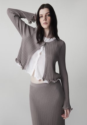 Vrouw poseert terwijl ze een taupekleurige geribbelde cardigan en rok draagt, gecombineerd met een wit, met knopen voorzien crop top, tegen een effen lichte achtergrond.