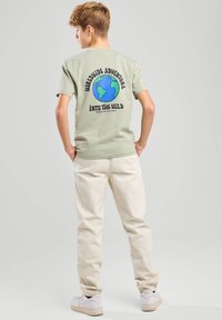 Camiseta de algodón verde con un gráfico en la parte posterior que representa un globo terráqueo y el texto "Aventura Mundial en la Naturaleza". Combinada con pantalones beige.