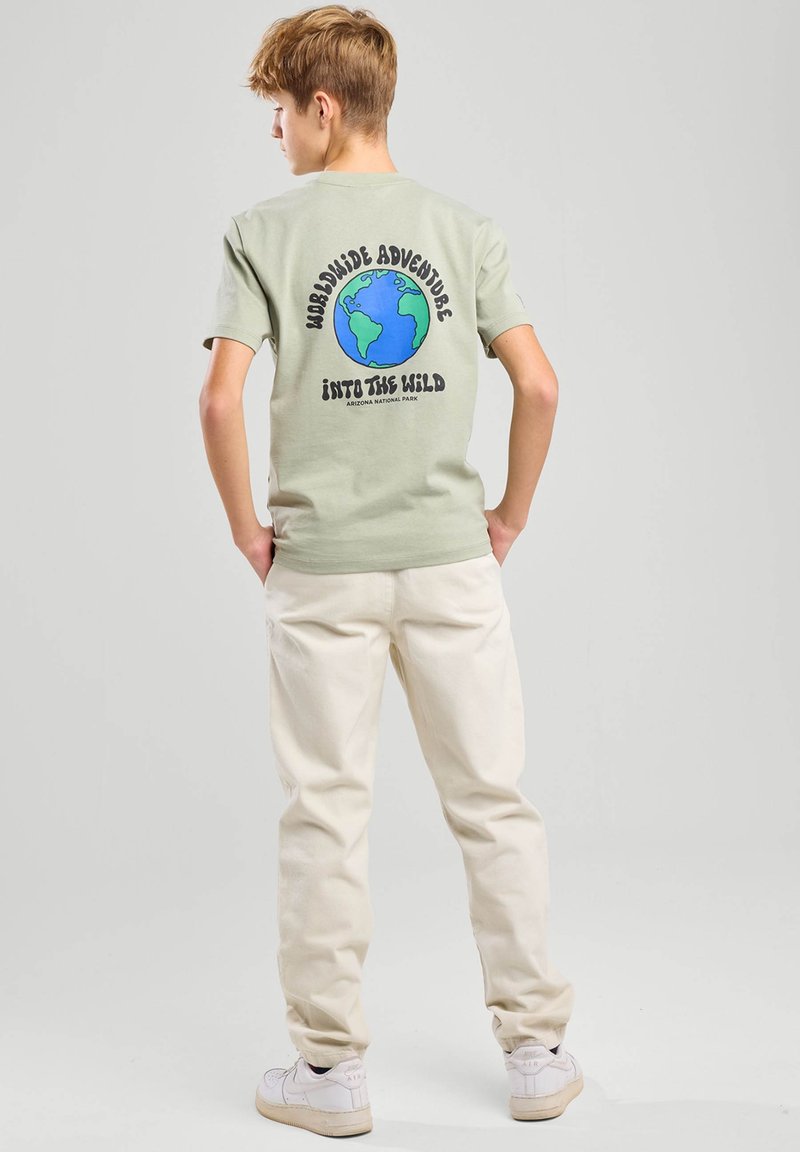 Teen boy standing with hands in pockets wearing light green t-shirt with globe and text "Worldwide Adventure Into The Wild" on back.