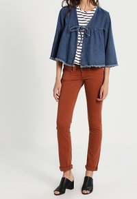 Farkkutakki, jossa on kuluneet reunat, nauhakiinnitys edessä, yhdistettynä raidalliseen toppiin, ruosteenvärisiin istuviin'housuihin ja mustiin korkokenkiin.