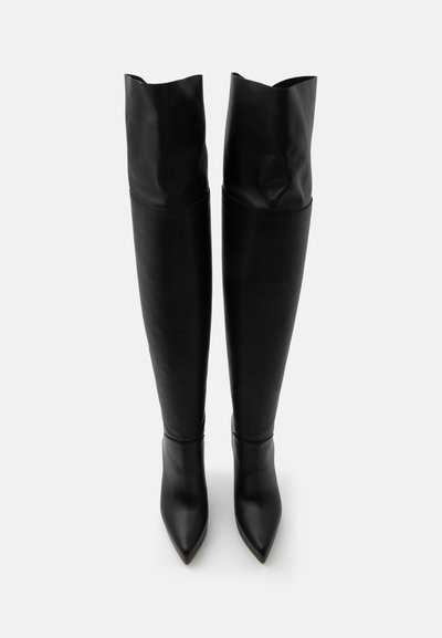 Bottes en cuir noires montantes jusqu'aux genoux avec des bouts pointus et des hauts amples, debout sur un fond blanc.