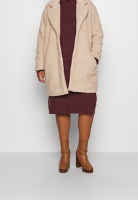 Beige teddyjacka med en mjuk, texturerad yta. Vinröd stickad klänning under. Modellen bär bruna klackade ankelskor med blockklack.