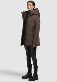 Brown padded jacket with a large hood, zipped front, and two side pockets. Worn with black fitted pants and black ankle boots.